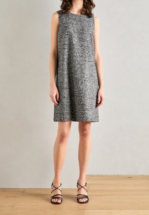 Kvinde iført en ærmeløs, knælængde grå tekstureret kjole og sorte sandaletter med stropper og høje hæle, stående på et trægulv.