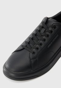 Sorte skinnesneaker med rund tå, teksturert gummisåle, seks snorløkker og flate skolisser; har preget logo på siden.