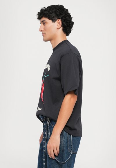 Čierne tričko s krátkym rukávom a grafickým motívom čerešne na prednej strane, v kombinácii s modrými džínsami so zlatým detailom na páse.