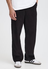 Pantaloni negri din catifea cu un design cu picioare largi, având o textură verticală cu dungi și buzunare laterale, asortați cu adidași albi și gri.