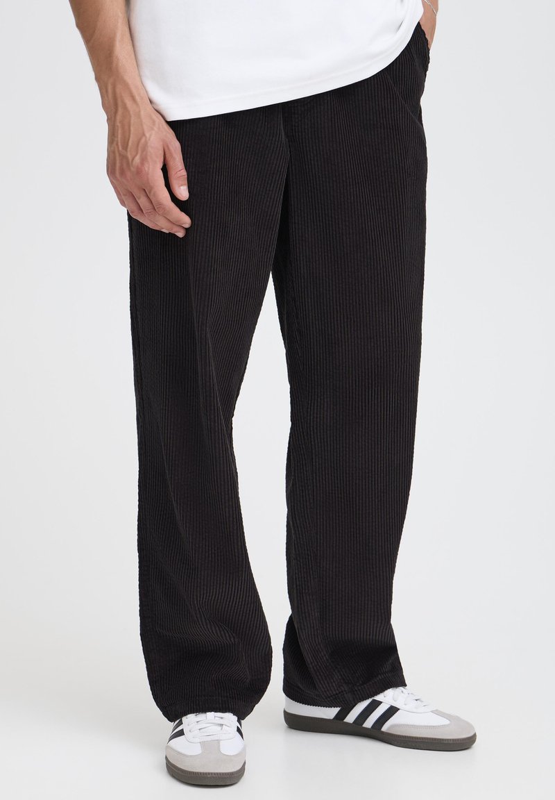 Pantaloni negri din catifea cu un design cu picioare largi, având o textură verticală cu dungi și buzunare laterale, asortați cu adidași albi și gri.