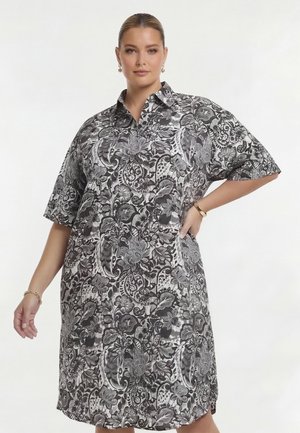 Kvinde iført en knælængde, kortærmet kjole med sorte og hvide blomster- og paisley-mønstre, poserer med den ene hånd på hoften.