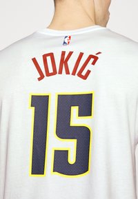 Maillot de basketball blanc avec le nom "JOKIĆ" en lettres rouges au-dessus du numéro "15" en bleu marine avec un contour jaune et une texture pointillée.