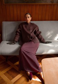 Brown oversized wool jacket with black buttons, paired with a flowing burgundy skirt and black leather gloves, seated on a light gray couch.