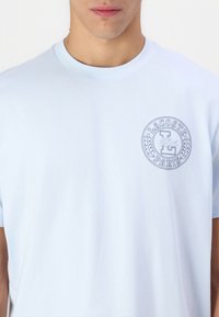 Svetlomodré bavlnené tričko s krátkymi rukávmi a okrúhlym výstrihom; obsahuje okrúhlu vyšitú nášivku s textom "LACOSTE" a "PARÍŽ".