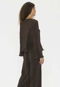 Donna con capelli ricci che indossa un top lungo a maniche pleated marrone scuro e pantaloni larghi abbinati, girata di lato su uno sfondo semplice.