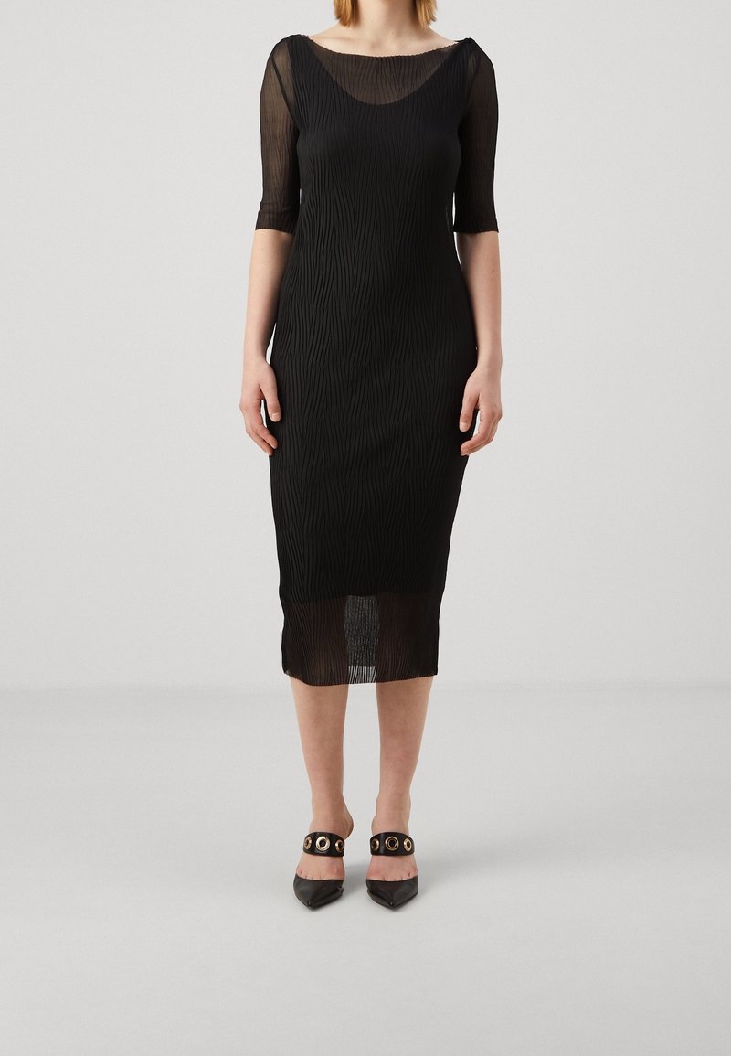 Femme portant une robe noire ajustée longueur genou avec manches mi-transparentes et ourlet transparent, associée à des escarpins noirs à bouts pointus avec œillets dorés.