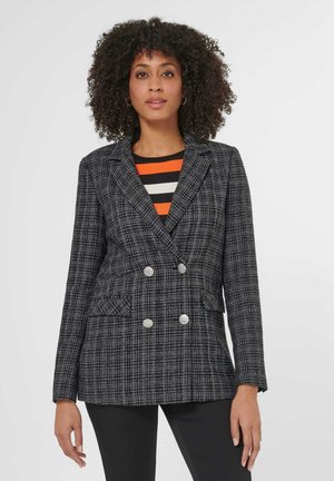 Frau mit lockigem Haar, die einen schwarz-weiß karierten, zweireihigen Blazer über einem gestreiften orange-, schwarz-weißen Oberteil trägt.