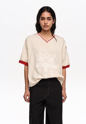 Młoda kobieta o długich ciemnych włosach nosi luźną beżową koszulkę z dekoltem w szpic ze czerwonym obszyciem oraz czarne spodnie na jednolitym białym tle.