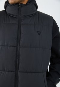 Gilet rembourré noir avec col haut, fermeture éclair à l'avant et accent logo. Présente une texture matelassée et pas de manches, porté par-dessus un sweat à capuche noir.
