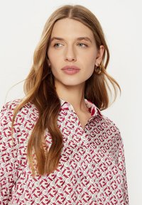 Camicia con base bianca caratterizzata da un motivo geometrico rosso e rosa. Design con bottoni, tessuto morbido e dettaglio del colletto classico.