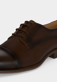 Chaussure de ville homme en cuir marron foncé avec bout droit, laçage fermé et petit talon en bois sur fond clair uni.
