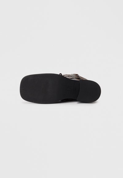 Black rubber sole of a sandal-style shoe with a textured finish, featuring leather straps and metallic hardware accents.