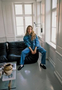 Veste et pantalon en jean bleu, canapé en cuir noir, table basse en verre avec des magazines, vase en céramique, grandes fenêtres aux cadres blancs.
