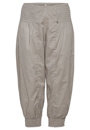 Ung person iført en hvid ærmeløs top, beige korte bukser med elastiske manchetter og hvide sneakers, stående mod en ensfarvet baggrund.