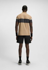 Kortærmet tan sportstrøje med mørkegrå stribe, kombineret med sorte shorts. Har logo på kraven og håndledsbånd, hvilket giver et strømlinet design.