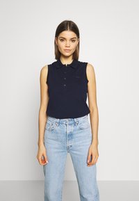 Temno modra brezrokavna polo majica z gumbastim zapenjanjem, rebrastim ovratnikom in logotipom Lacoste; v kombinaciji s svetlo modrimi visokimi jeans hlačami.
