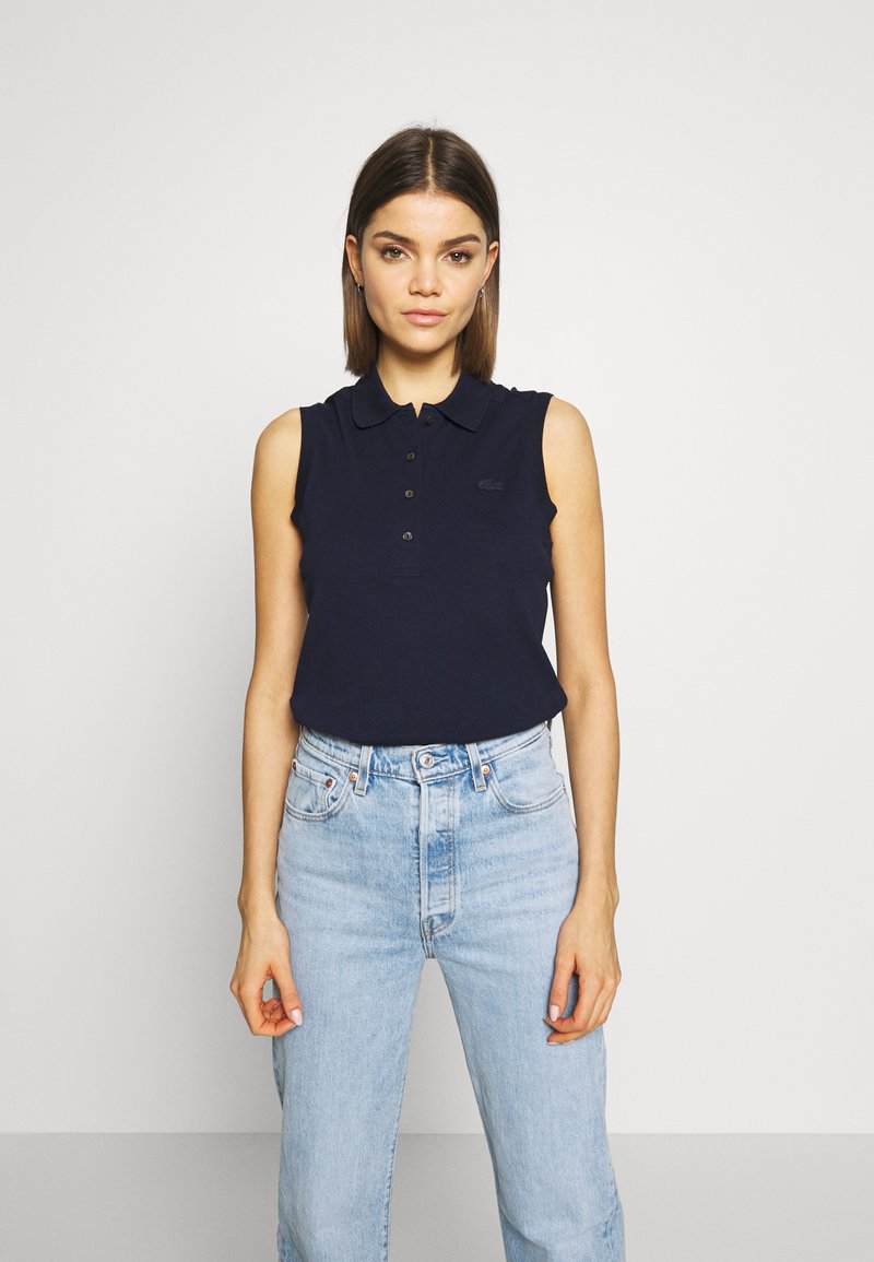 Temno modra brezrokavna polo majica z gumbastim zapenjanjem, rebrastim ovratnikom in logotipom Lacoste; v kombinaciji s svetlo modrimi visokimi jeans hlačami.