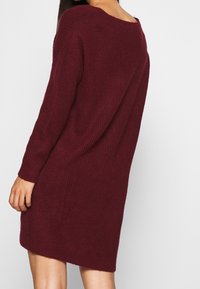 Robe pull tricotée bordeaux avec une coupe ample, des manches longues et un col large, présentant une texture côtelée et une longueur au-dessus du genou.