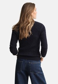 Marineblauer Pullover mit Zopfmuster, langen Ärmeln und einem gerippten Saum, kombiniert mit locker sitzenden Jeans und einem kontrastierenden weißen Hemdärmel.