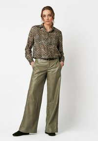 Woman wearing a sheer leopard print blouse tucked into high-waisted olive green wide-leg pants with hands in pockets.