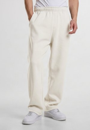 Homme portant un pantalon de survêtement ample beige clair avec une taille élastique et des baskets blanches sur un fond clair uni.