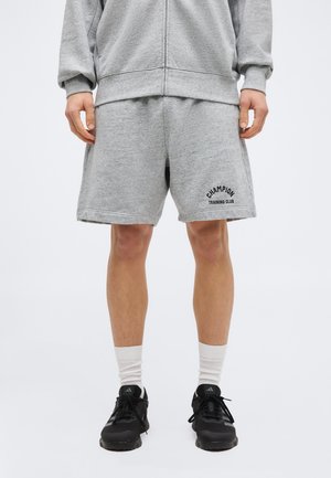 Άτομο που φορά γκρι σορτς Champion Training Club, γκρι φούτερ, λευκές κάλτσες και μαύρα αθλητικά παπούτσια Adidas, στέκεται σε απλό φόντο.