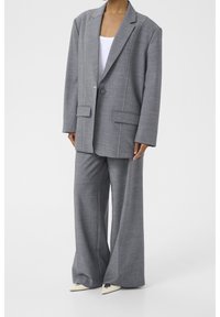 Harmaa ylikokoinen puku, jossa on räätälöity bleiseri ja leveälahkeiset housut, joissa on kontrastivaadit ja kaksi etutaskua.