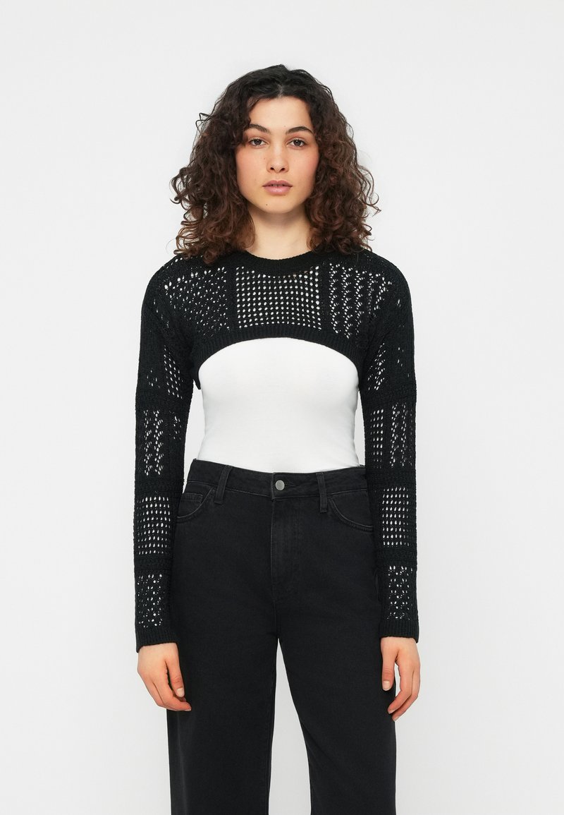 Γυναίκα με σγουρά μαλλιά φοράει μαύρο ανοιχτό πλεκτό cropped πουλόβερ πάνω από λευκό τοπ και μαύρο παντελόνι ψηλόμεσο, στέκεται μπροστά σε απλό φόντο.