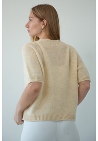 Jasno beżowy sweter dziany z krótkim rękawem, z prążkowanymi mankietami i szerokim dekoltem. Materiał ma miękką, puszystą fakturę.