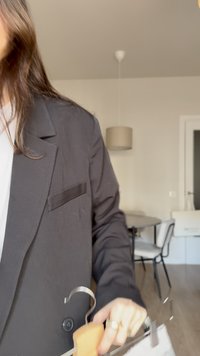 Person trägt einen schwarzen Blazer und hält einen Holzkleiderbügel mit einem weißen Kleidungsstück in einem hellen Raum mit einem runden Tisch und Stühlen.