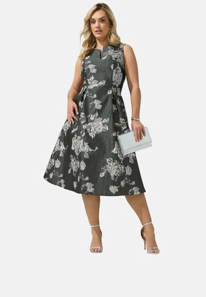 Kvinde iført sort midi-kjole uden ærmer med sølvfarvet blomsterprint, holdende en sølvfarvet clutch og iført sølvfarvede højhælede sandaler.