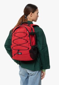 Sac à dos rouge en tissu texturé, avec un cordon élastique à l'avant, des sangles ajustables et des accents en maille respirante. Caractérisé par le logo de la marque.