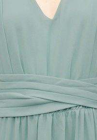 Κοντινή λήψη υφάσματος ανοιχτού γαλάζιου φορέματος με V-λαιμόκοψη και λεπτομέρεια με σούρες στη μέση που δημιουργεί απαλές πτυχές.