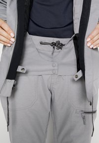 Mains ajustant le devant ouvert d’un pantalon gris à taille avec cordon, boutons-pression et poche zippée sur la cuisse droite.