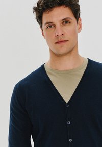 Granatowy kardigan z dekoltem w serek, wykonany z miękkiej tkaniny. Posiada pięć guzików z przodu i jest nakładany na jasnobeżową koszulę.