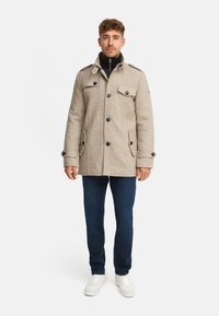 Manteau beige en mélange de laine avec un col montant, des poches avant et une fermeture à boutons. Associé à un jean foncé et des baskets blanches.