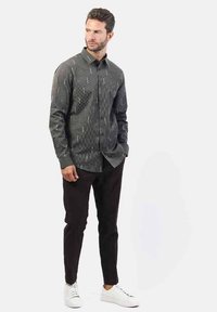 Camicia a maniche lunghe a fantasia bianca e nera, abbinata a pantaloni scuri e sneakers bianche, in piedi su uno sfondo neutro.