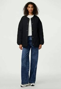 Chaqueta negra acolchada con botones a presión, llevada sobre una camiseta blanca, combinada con unos jeans azul de cintura alta y zapatillas blancas.