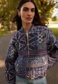 Chaqueta bomber acolchada en tonos morados con patrones geométricos y florales, cremallera de metal y cuello alto. Tejido ligero con textura.