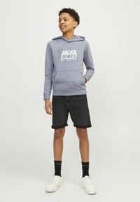 Sweatshirt à capuche gris avec logo blanc, associé à un short en jean noir. Porté avec des baskets blanches et des chaussettes noires. Style décontracté.