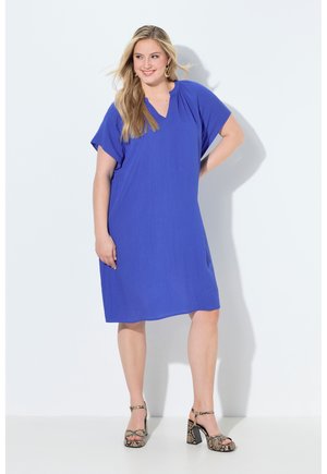 Uśmiechnięta kobieta o długich blond włosach, ubrana w luźną niebieską sukienkę do kolan i sandały na wysokim obcasie w wężowy wzór, stojąca na białym tle.