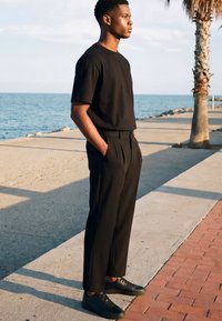 Jeune homme en tenue noire debout, les mains dans les poches, sur un chemin au bord de la mer et d'un palmier en journée.