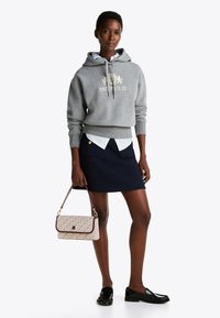Felpa grigia con cappuccio e logo, indossata sopra una camicia bianca, abbinata a una gonna blu navy. Tiene una borsa con motivo e indossa mocassini neri.