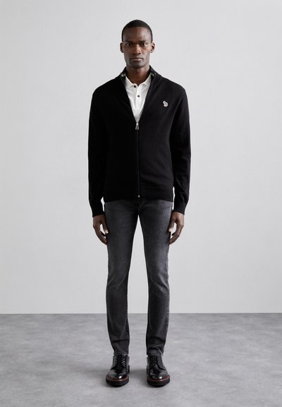 Black zip-up cardigan with a knitted texture, contrasting collar stripe, worn over a white polo shirt, paired with slim-fit gray jeans and black shoes.