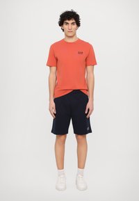Rød bomull T-skjorte med trykt logo, parret med marineblå shorts. Hvite sneakers og lysegrå sokker fullfører antrekket.