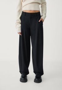 Pantalon noir à jambes larges en tissu lisse avec plis avant, poches latérales et taille ajustée, associé à des chaussures noires épaisses.