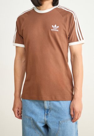 T-shirt en coton marron avec des accents blancs, présentant un col rond, des manches raglan courtes, et un petit logo Adidas blanc sur la poitrine.