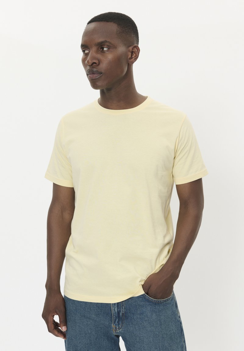 Camiseta de manga corta y cuello redondo en suave tela de color amarillo pálido. Presenta un corte relajado y un diseño sencillo, combinada con jeans de denim azul.