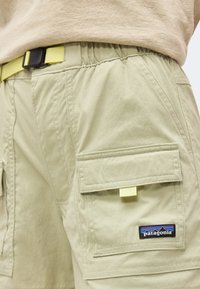 Μπεζ cargo σόρτς με μεγάλη πλαϊνή τσέπη και λογότυπο Patagonia, φορεμένα με μπεζ τοπ και στερεωμένα με κίτρινη ζώνη με μαύρη αγκράφα.